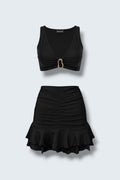 The Saint 54 black two-piece set featuring a fitted crop top with gold hardware and a ruched mini skirt with ruffles.