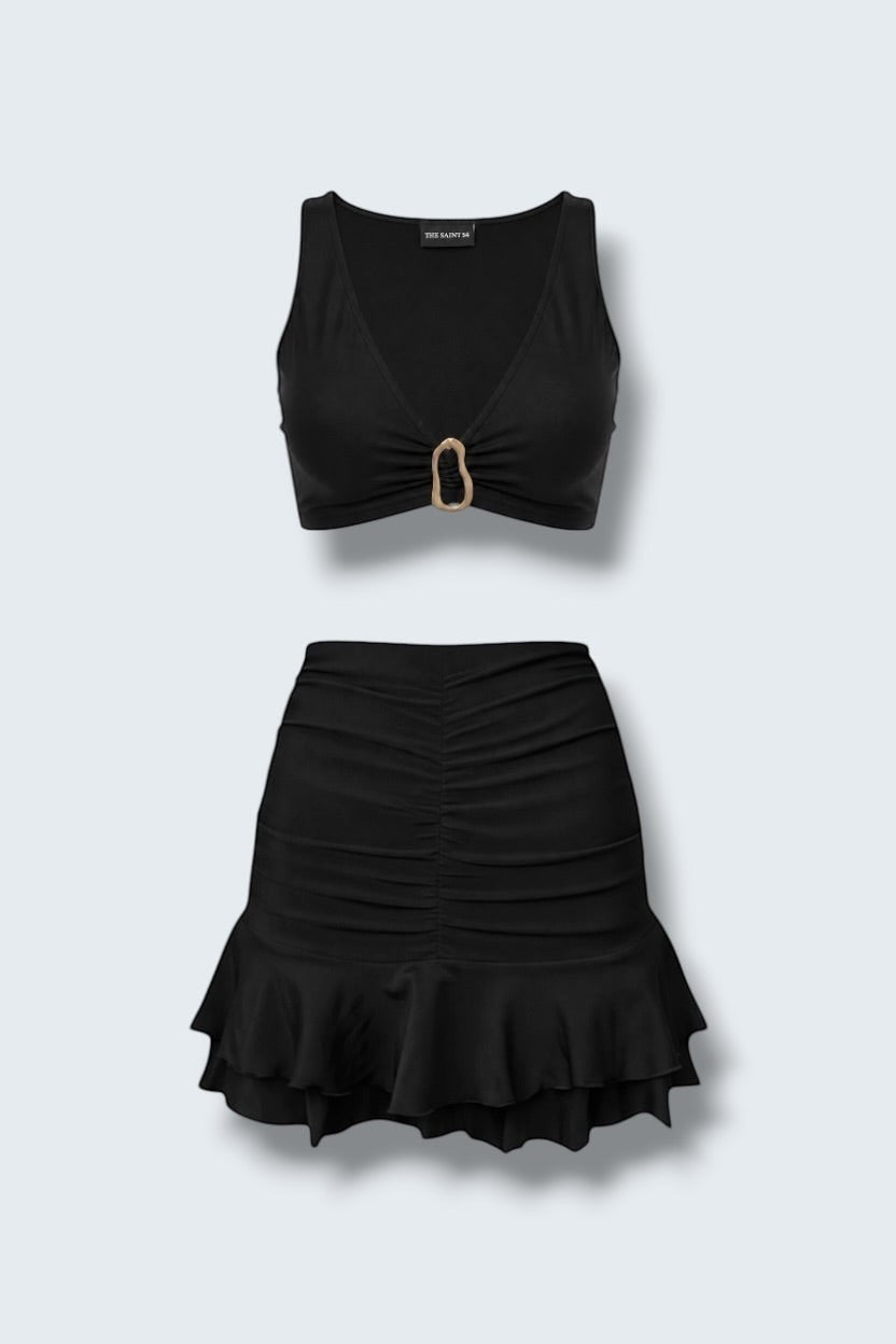 The Saint 54 black two-piece set featuring a fitted crop top with gold hardware and a ruched mini skirt with ruffles.
