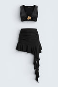 The Saint 54 black two-piece set featuring a fitted crop top with gold accent and an asymmetrical ruffled mini skirt.